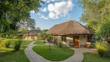 A thatched-roof bungalow with a stone pathway leading to the entrance, surrounded by lush greenery and trees.
