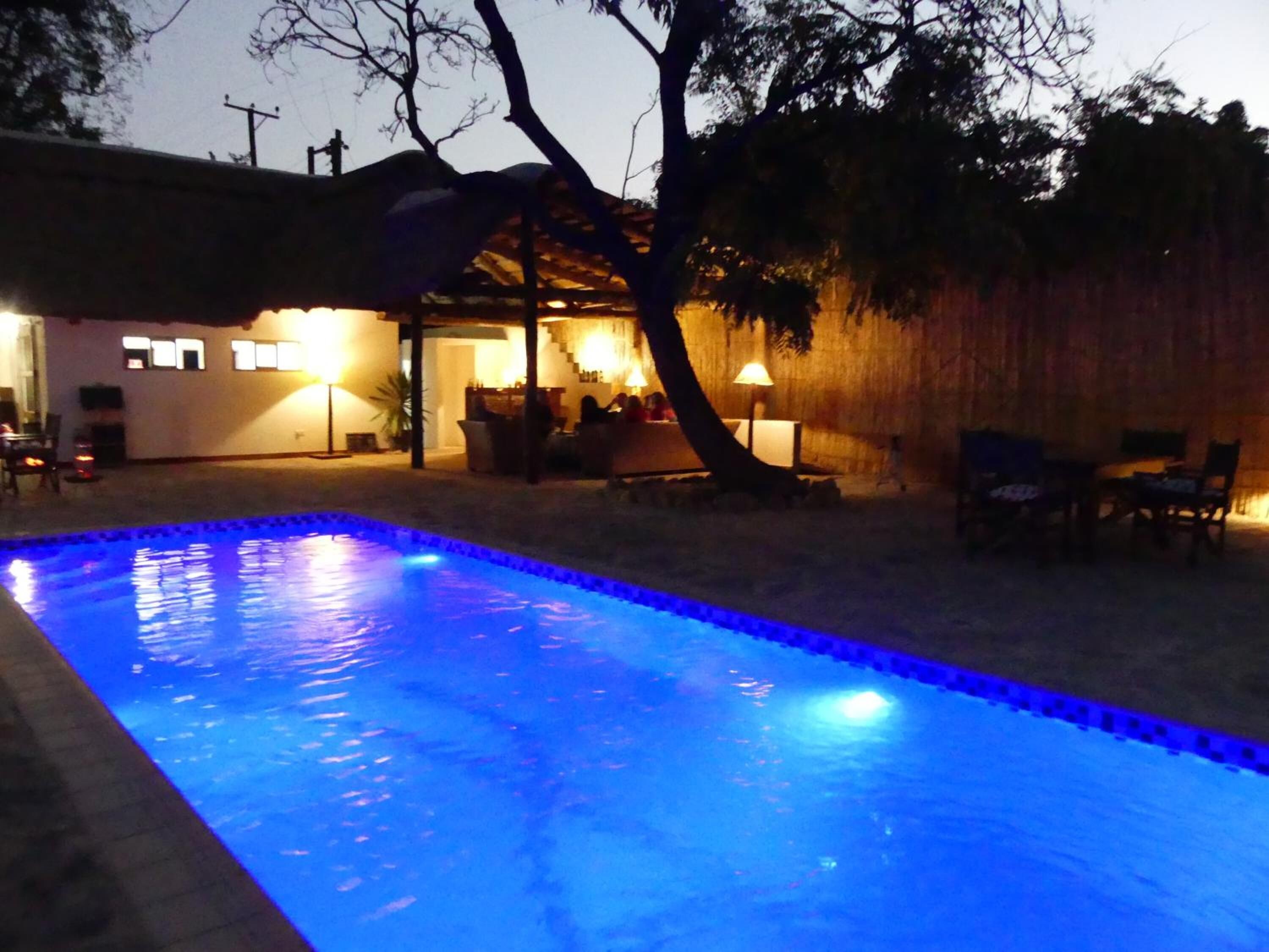 A swimming pool with a blue-lit edge, surrounded by outdoor furniture and a covered seating area with ambient lighting.