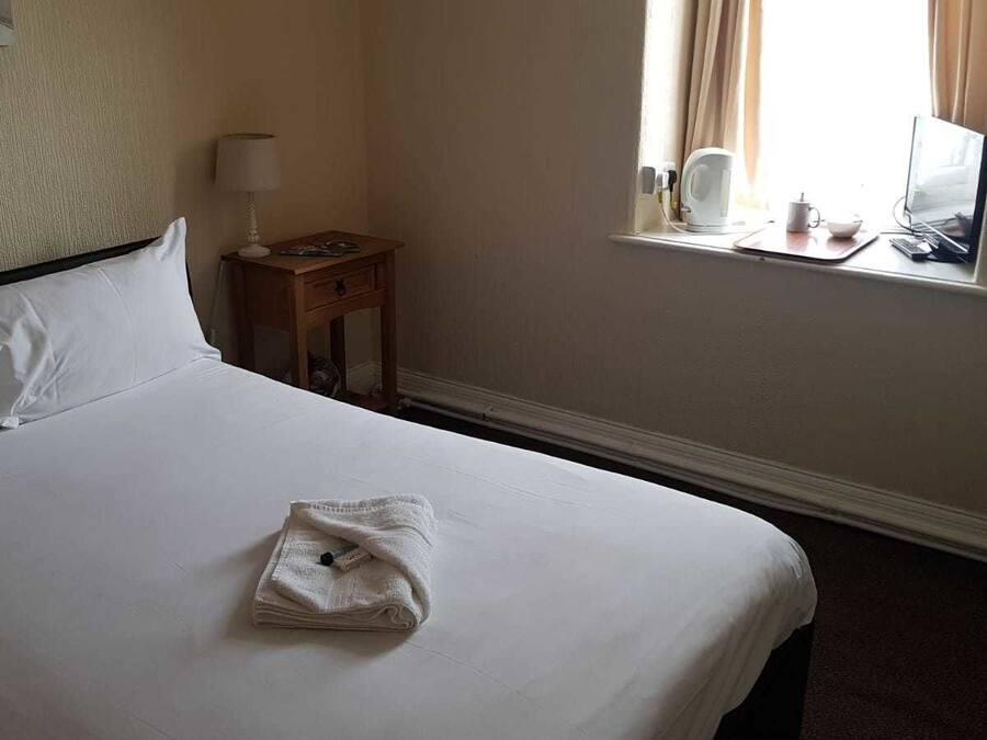 A neatly made bed with a folded towel, a bedside table with a lamp, and a window with curtains.