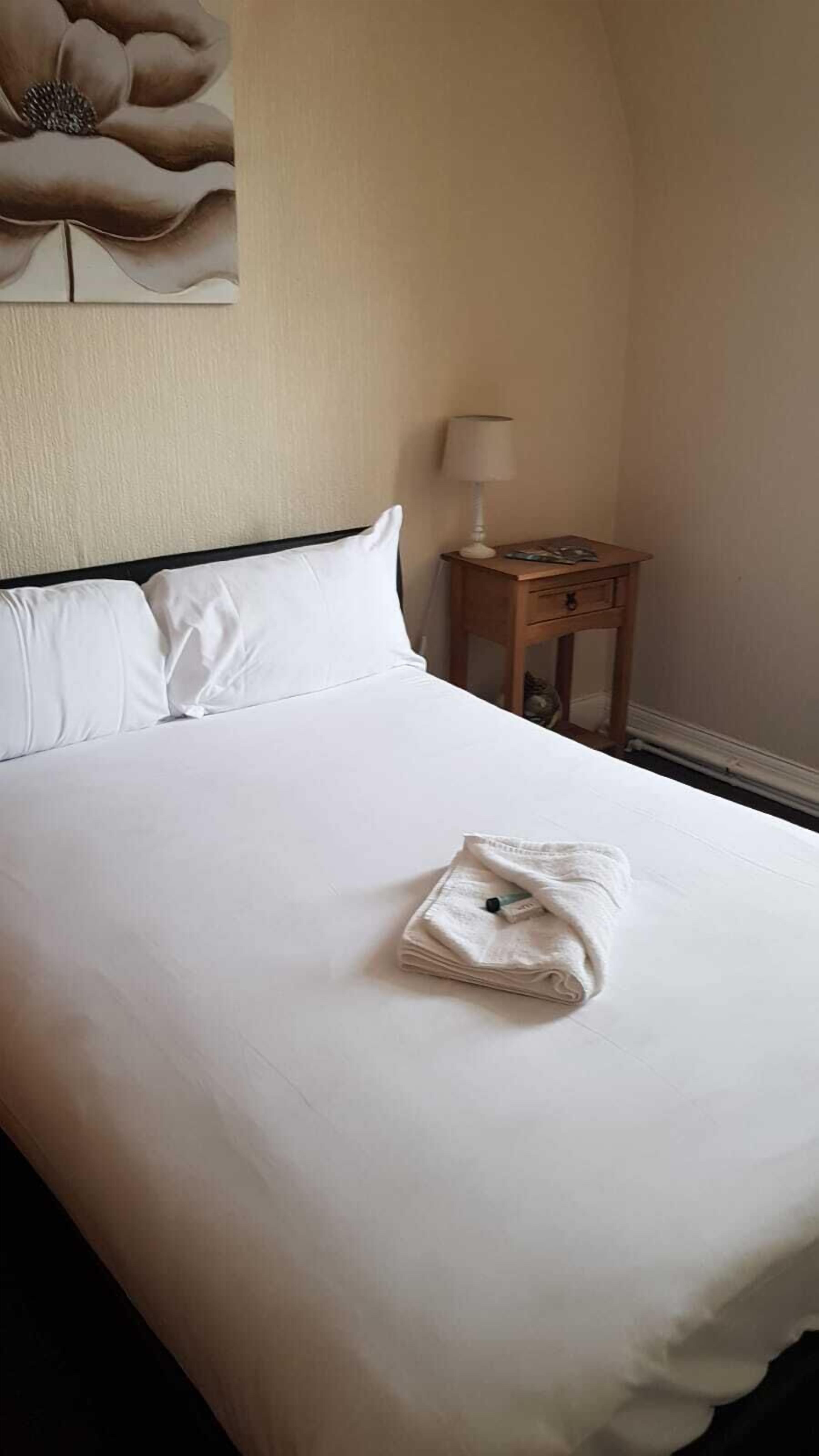 A neatly made bed with white linens, a folded towel on the bed, and a small wooden nightstand with a lamp beside it.