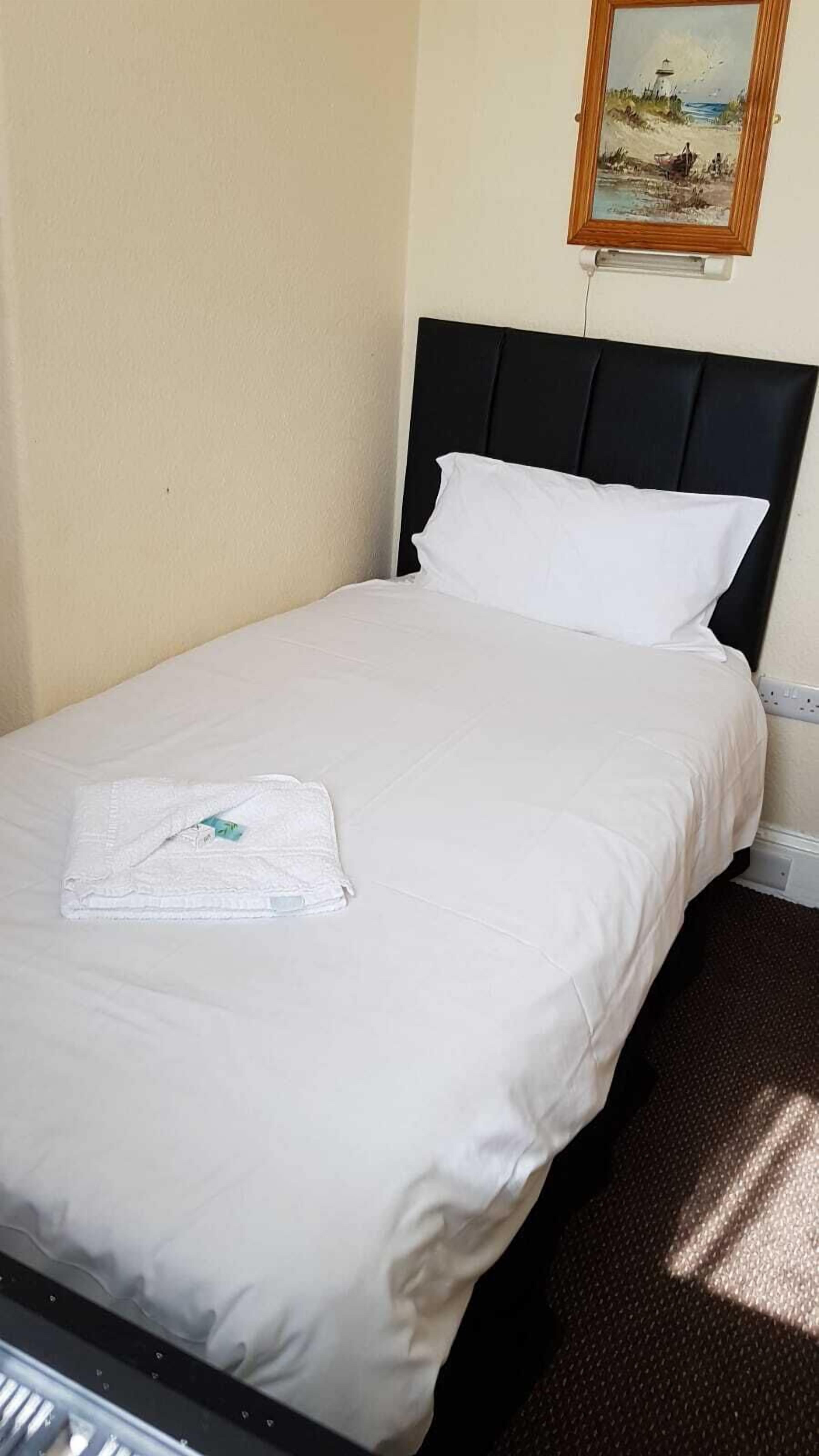 A neatly made bed with white linens and a folded towel, a black headboard, and a painting of a beach scene on the wall.