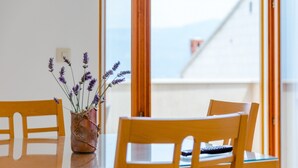Uma mesa de jantar com um vaso de flores de lavanda, cadeiras de madeira e vista para edifícios através de grandes janelas.