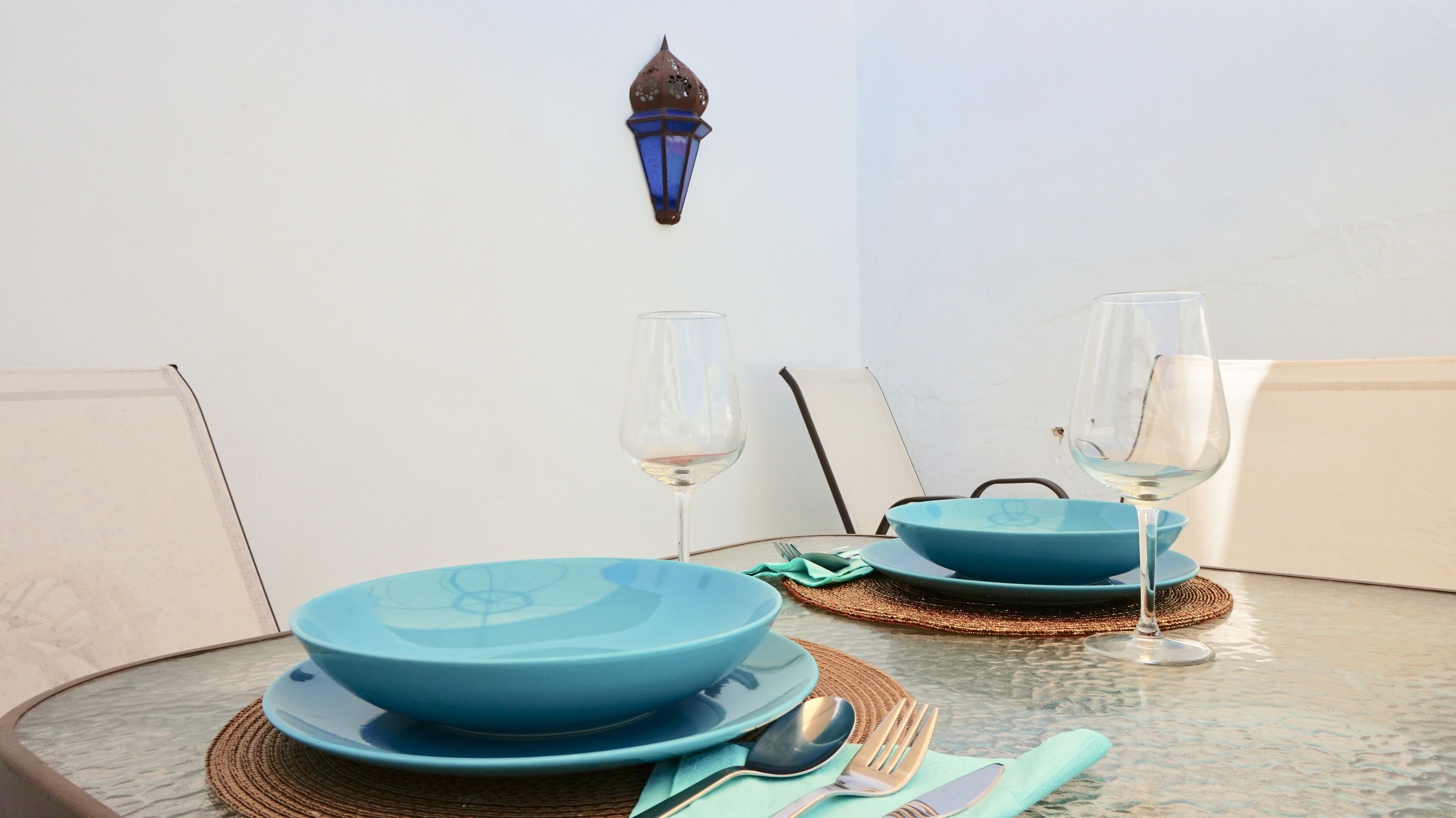 Une table à manger dressée avec de l’assiette bleue, deux verres à vin et des couverts, et une applique murale décorative au-dessus.