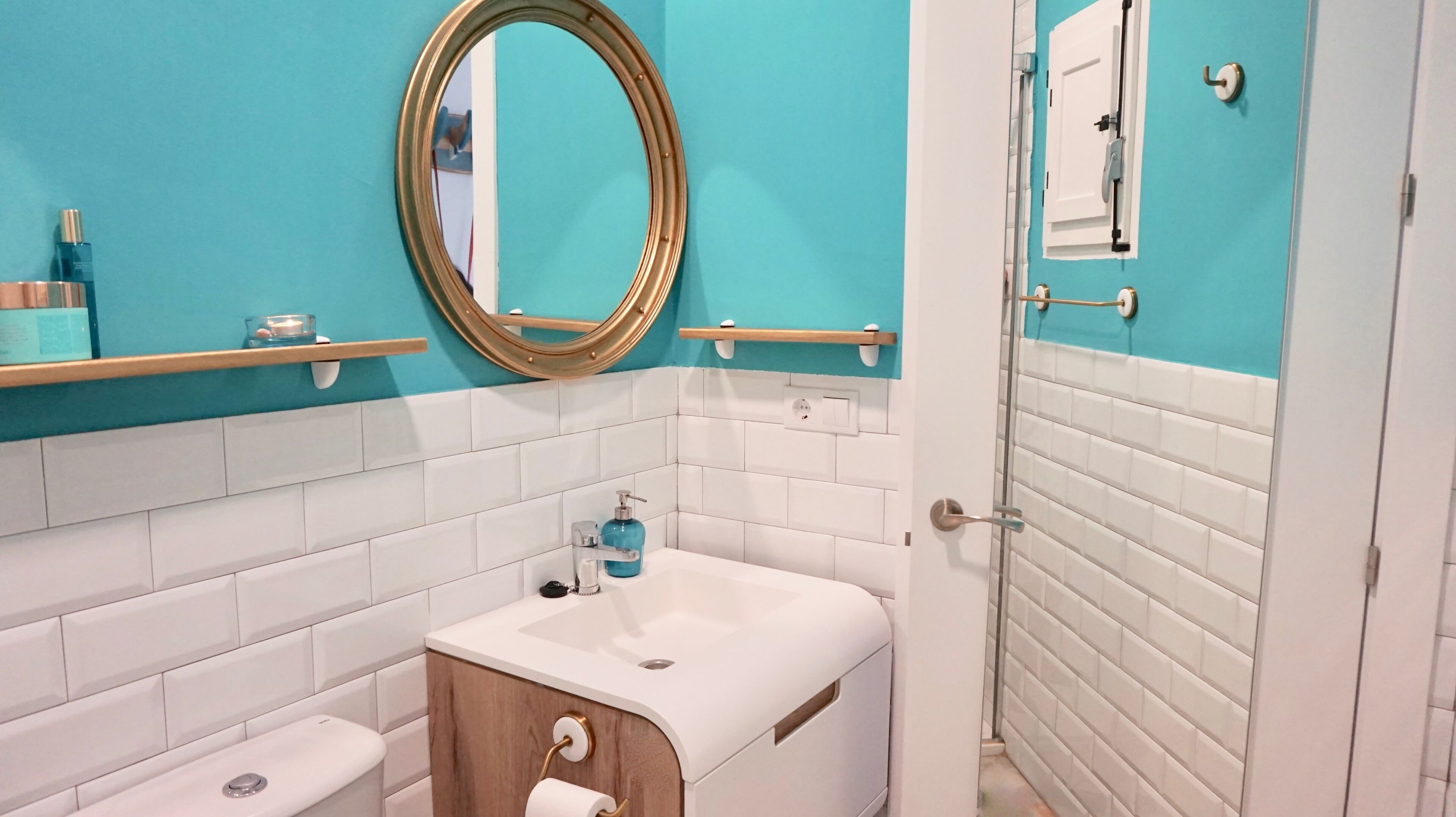 Casa de banho com lavatório branco, espelho e sanita. Encontra-se uma estante com artigos de higiene pessoal e um porta-toalhas.