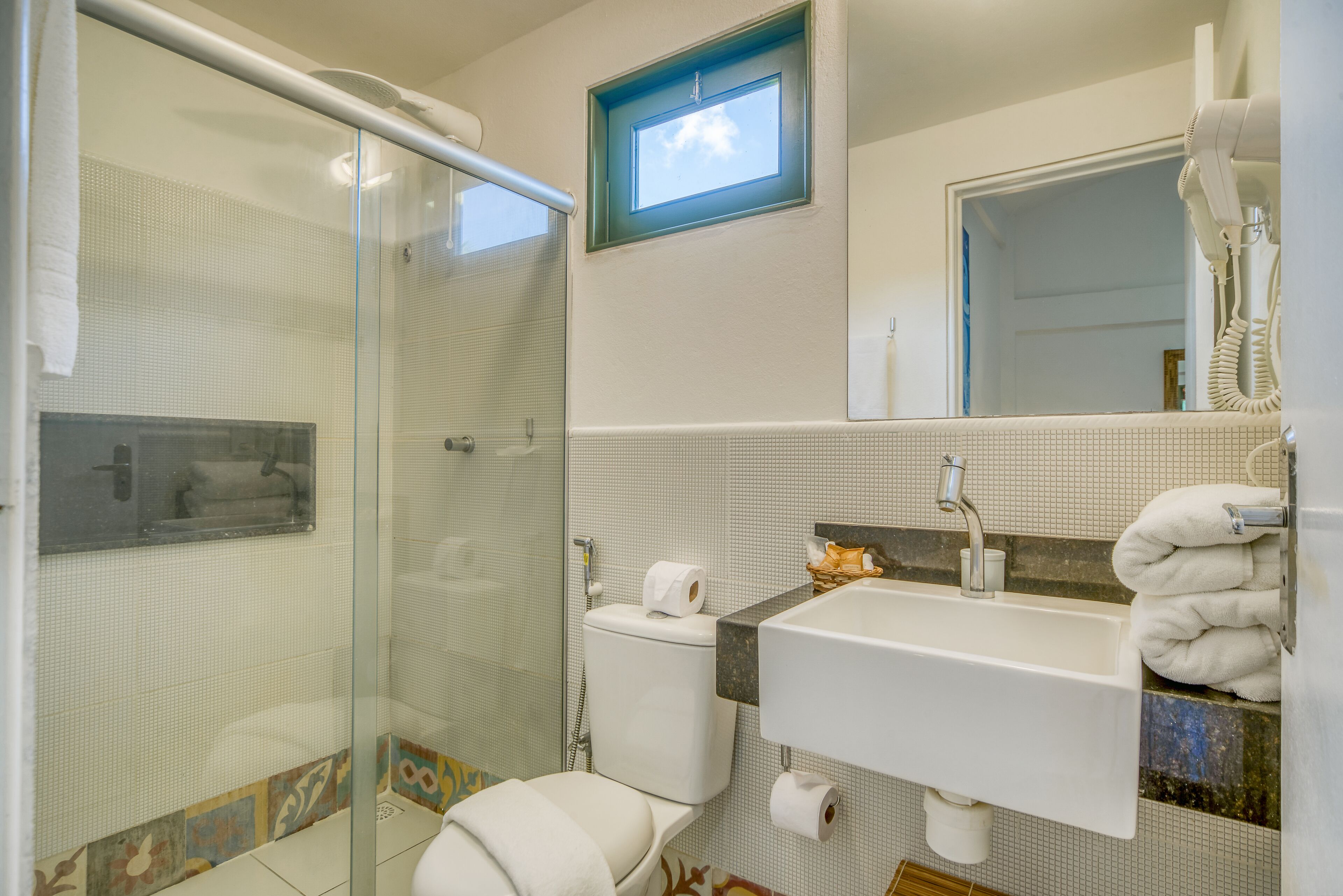 A bathroom with a glass shower enclosure, a toilet, a sink with a towel rack, and a small window.