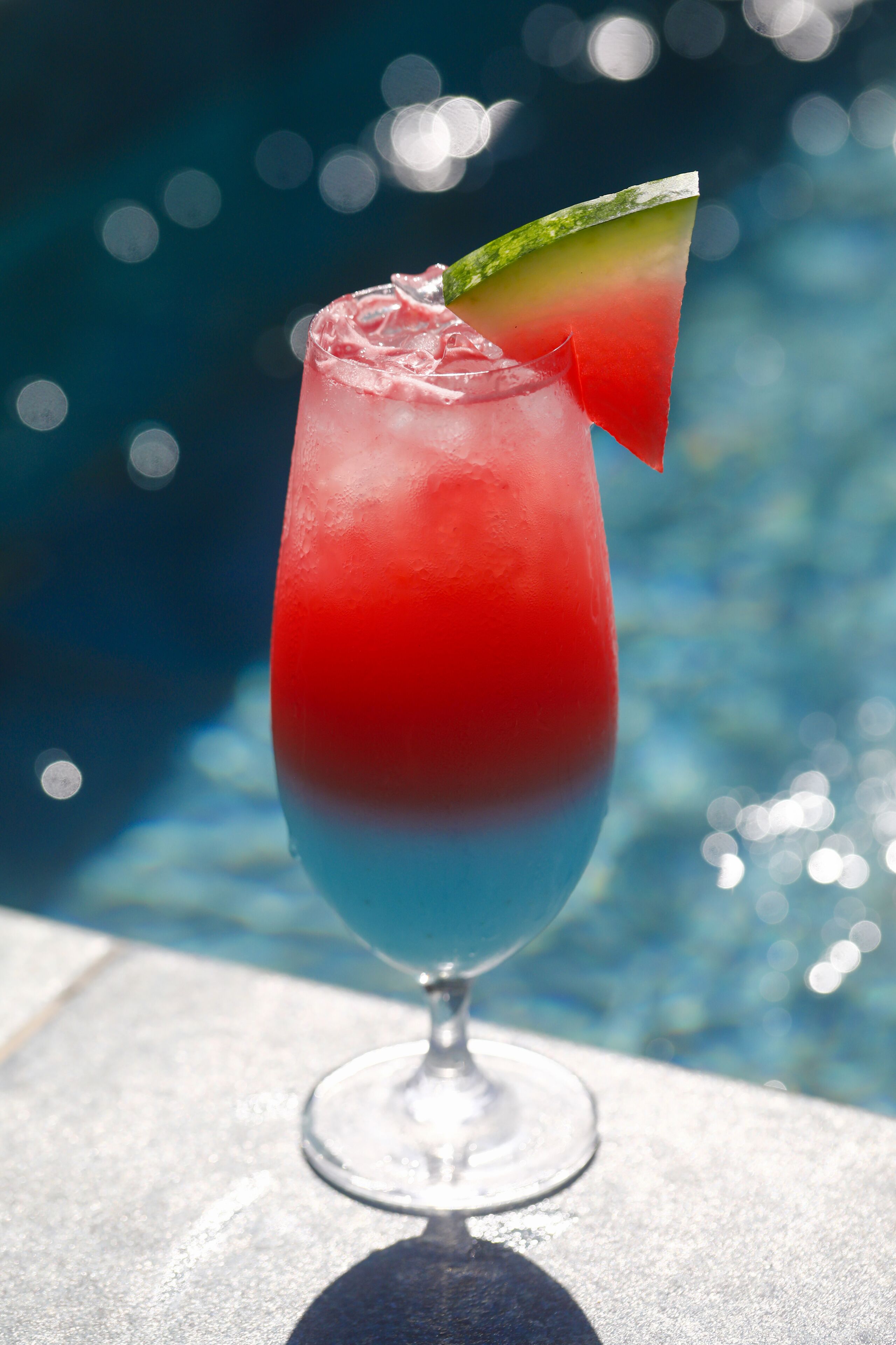 A cocktail glass with a red and blue gradient, garnished with a slice of lime, placed on a reflective surface near a pool.