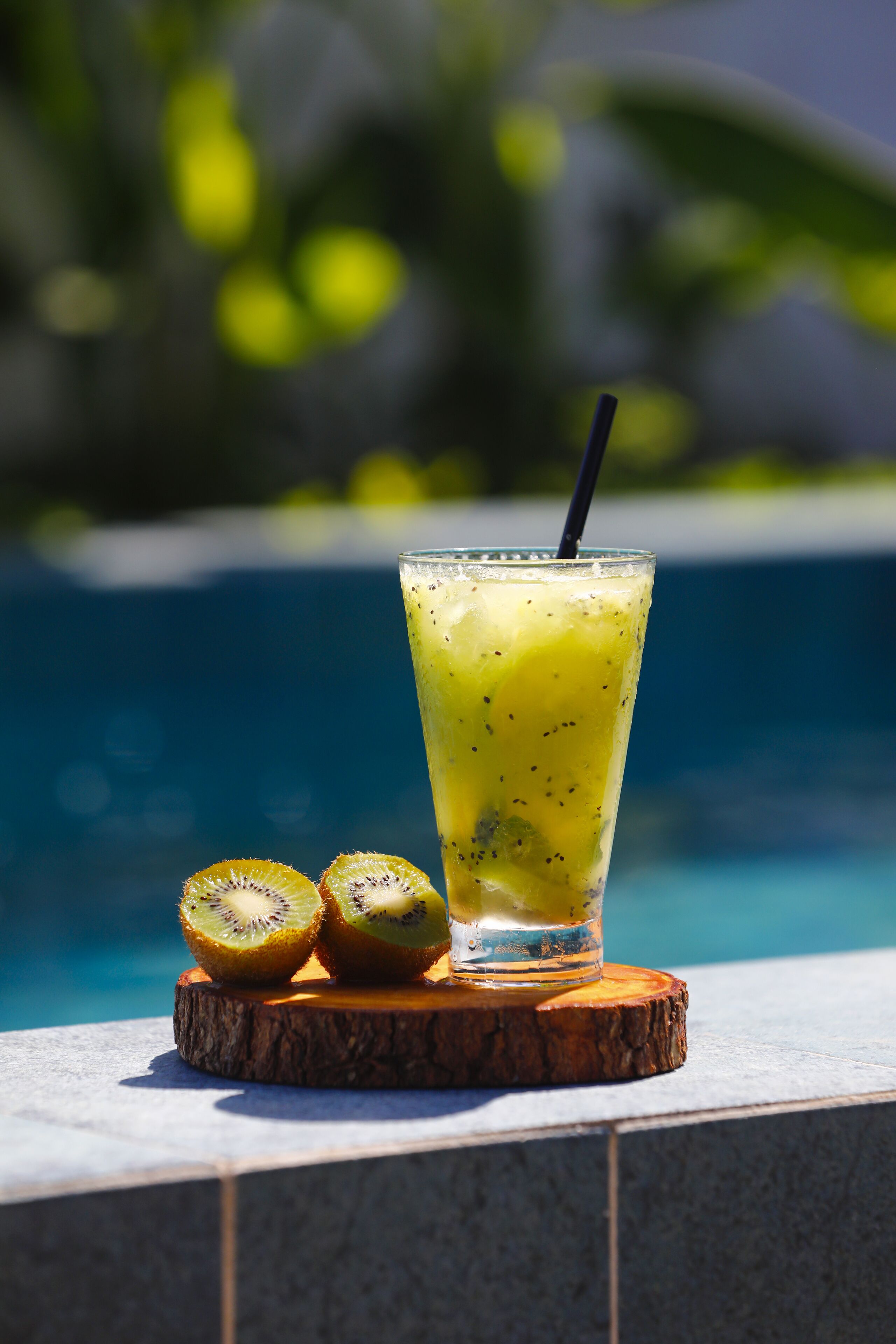 Un vaso de batido de frutas con pajita, sobre un posavasos de madera junto a una piscina.