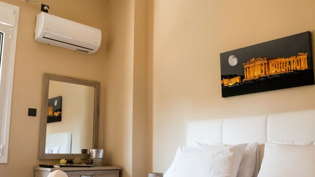 A hotel room with a bed, a bedside table, a mirror, an air conditioning unit, and a painting of a building. - Gazi Divine Apartment (Athens)