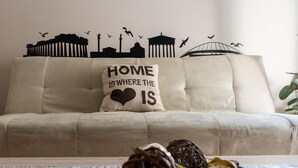 A beige sofa with a cushion that reads "HOME IS WHERE THE HEART IS" and a silhouette wall art depicting buildings and birds. - Gazi Divine Apartment (Athens)