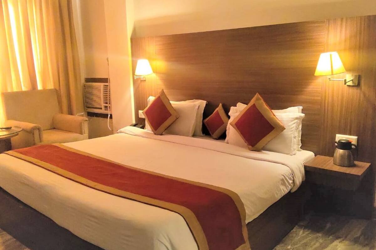 Habitación de hotel con una cama grande, dos lámparas de noche, una silla, una mesita pequeña y un calentador fijado a la pared.