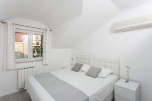 Un dormitorio moderno con una cama grande, ropa de cama blanca, una mesita de noche con lámpara y una ventana con cortinas.