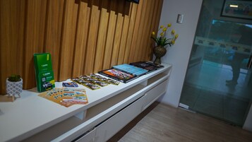 A reception desk with a white counter, a wooden slat wall, a vase with yellow flowers, and a glass door reflecting a person.