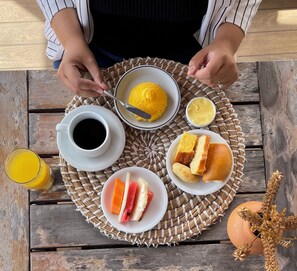 Una escena de desayuno con un posavasos tejido, una taza de café, un bol de gachas amarillas, un plato con pan y fruta, y un vaso de zumo de naranja.