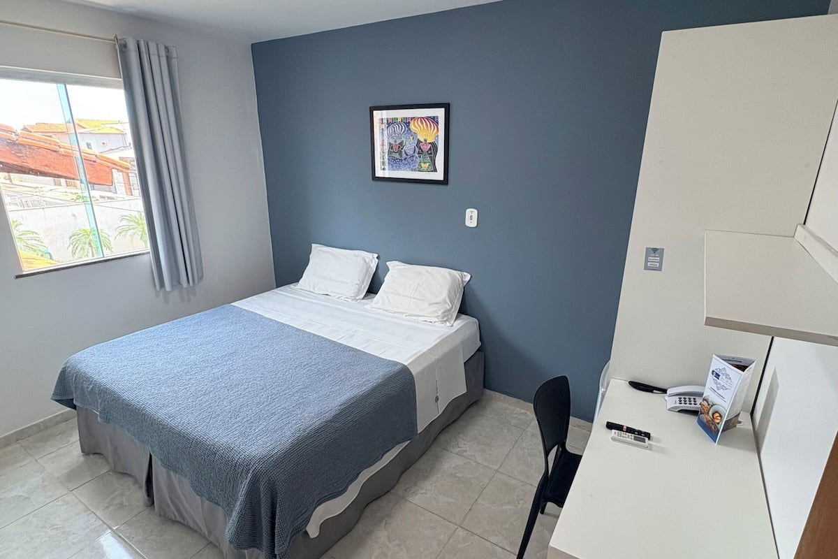 Quarto de hotel com cama, escrivaninha, cadeira, telefone e janela com cortinas.