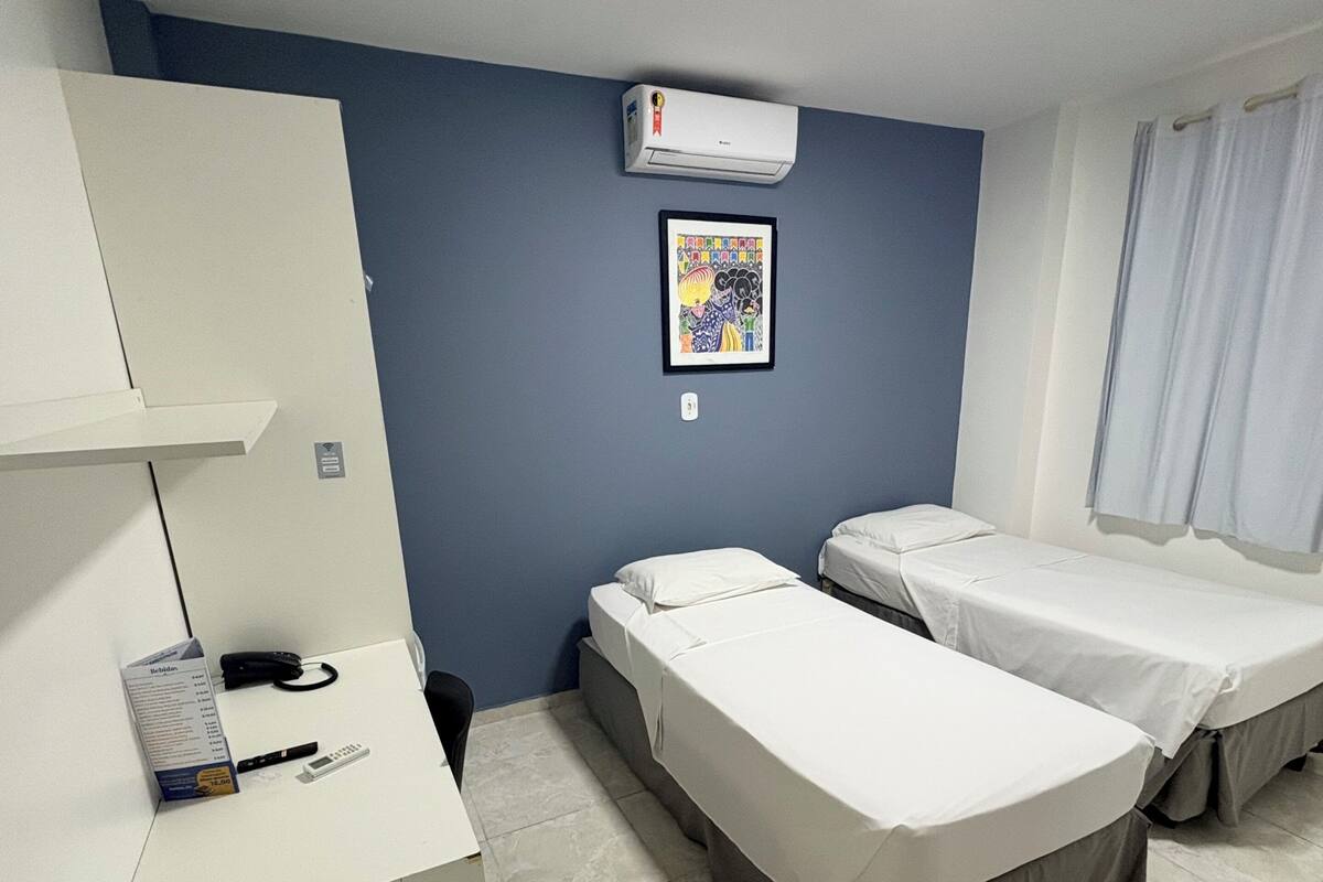 Quarto de hotel com uma mesa, duas camas, uma lixeira, ar condicionado e um quadro na parede.