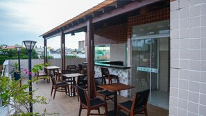 Un patio exterior con mesas y sillas de madera, un muro de ladrillo y una puerta de cristal que da a una zona de cocina.