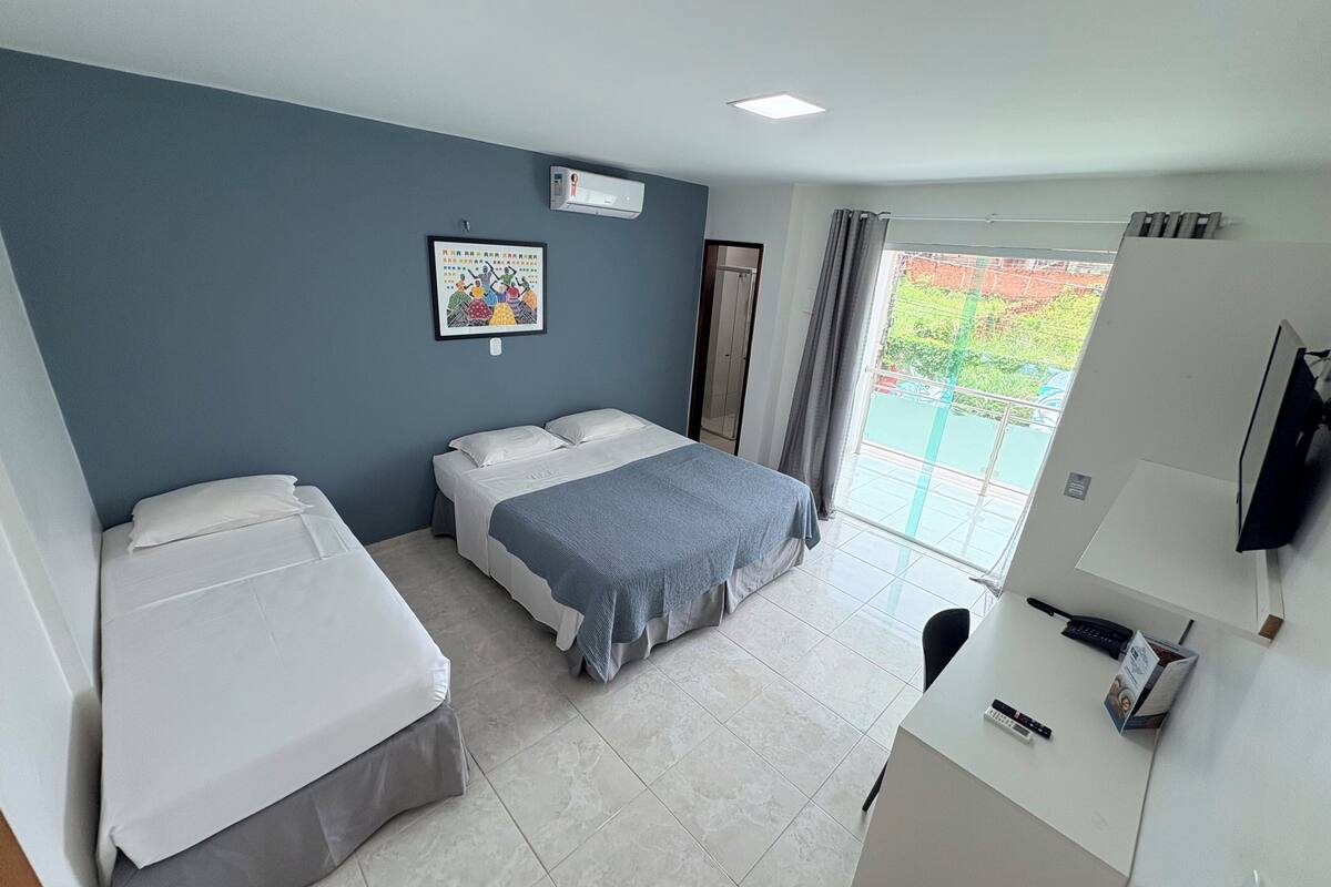 Quarto de hotel com duas camas, uma escrivaninha com telefone e controle remoto, uma televisão, uma janela com vista para o verde e um aparelho de ar condicionado na parede.
