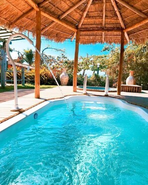 A swimming pool with a thatched roof structure, surrounded by palm trees and a clear sky. - Pousada Axé (Prado)