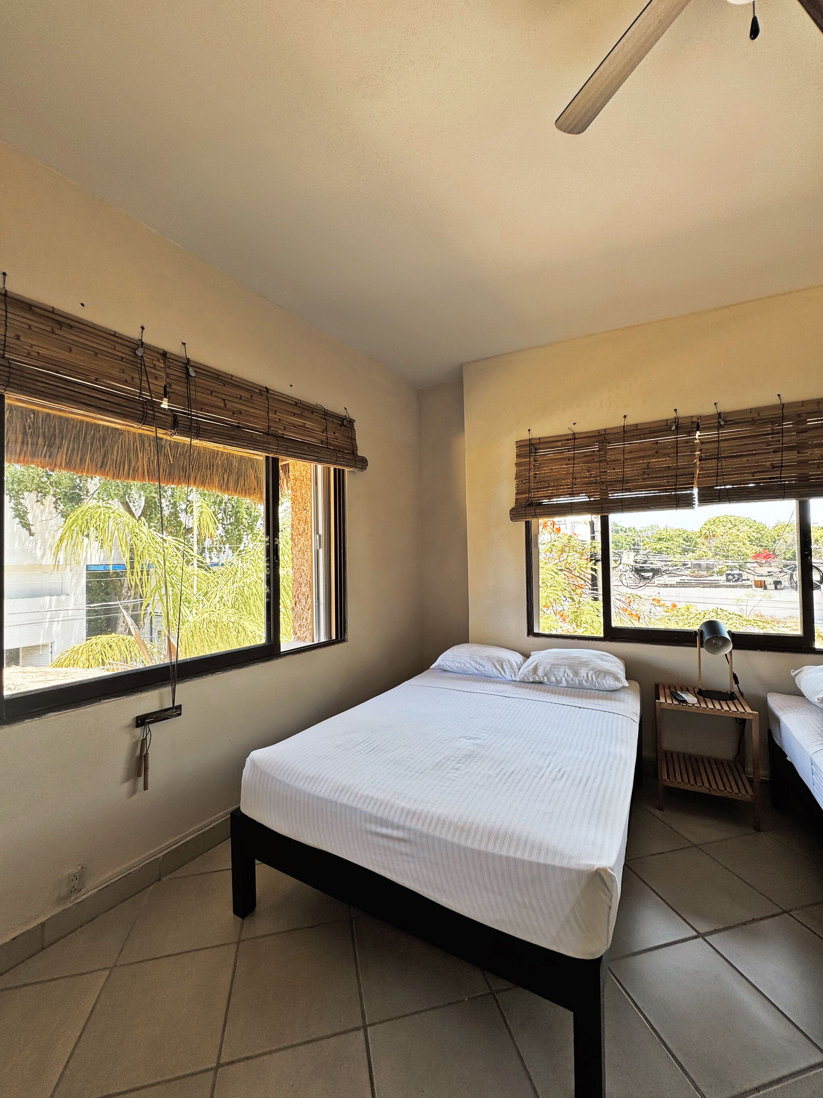 Un dormitorio con una cama, dos mesitas de noche y ventanas grandes con persianas.