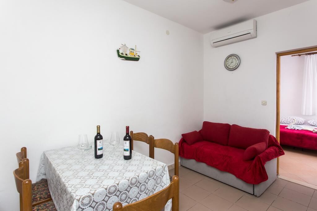 Una habitación pequeña con una mesa de comedor preparada para dos personas, un sofá rojo, un reloj y una pequeña decoración de barco en la pared.