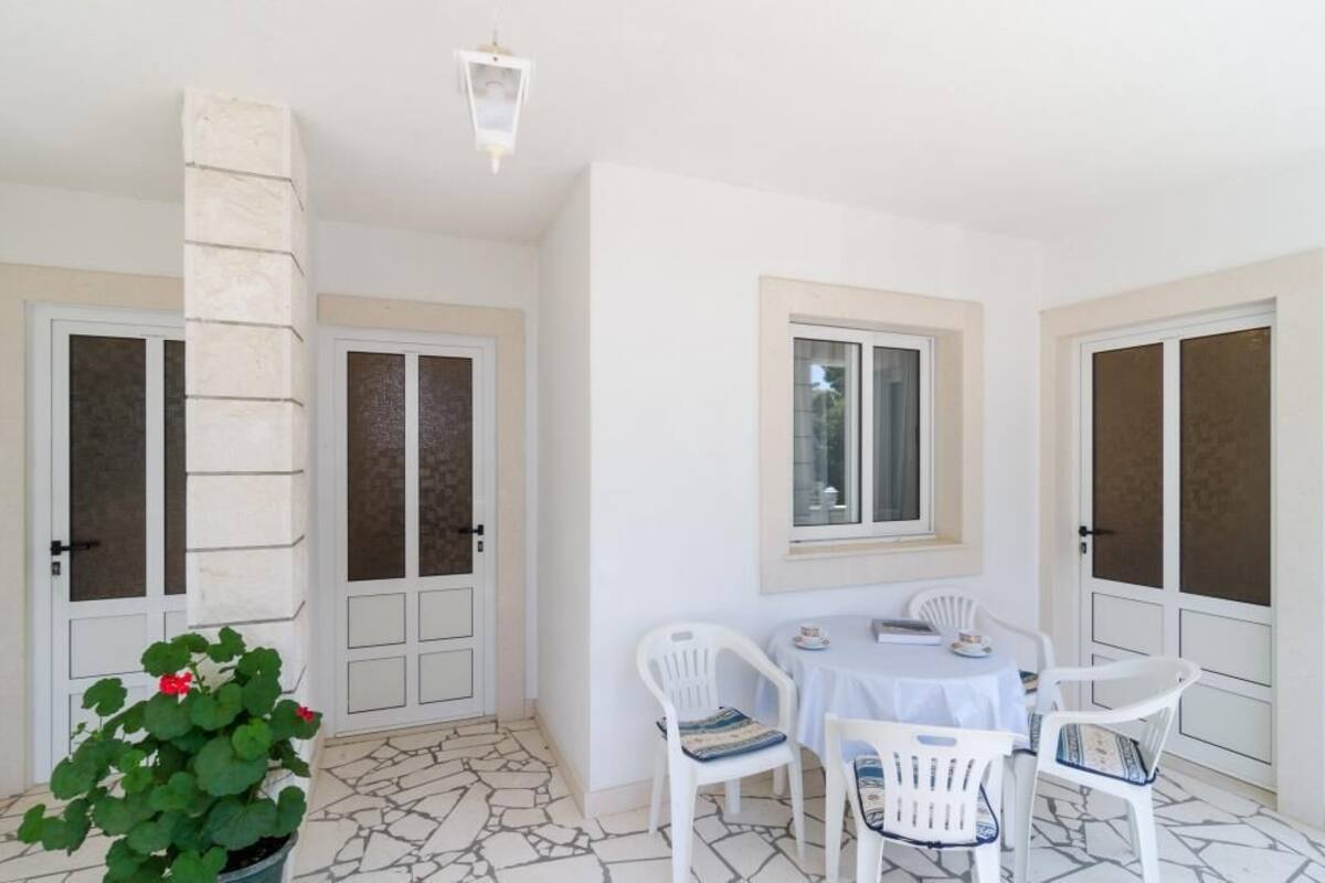 Una habitación con suelo de piedra, paredes blancas y una mesa redonda con cuatro sillas. Hay una ventana que ofrece vistas al exterior y una planta en maceta con hojas verdes y flores rojas.