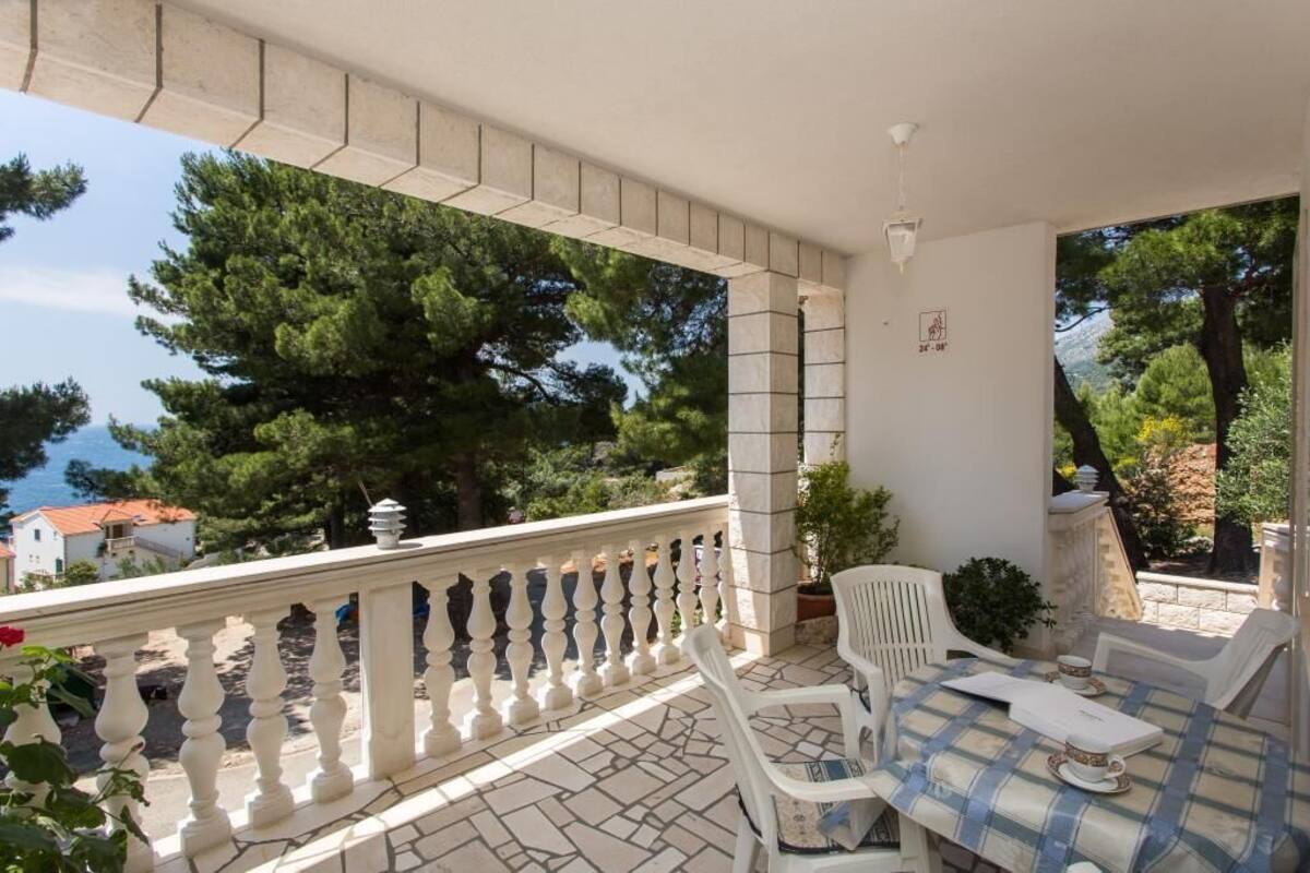 Un balcón con barandilla blanca, una mesa con sillas y vista al mar y a los árboles.