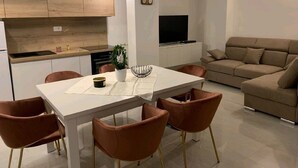 Un salon moderne avec une table à manger blanche, des chaises marron, un canapé beige et un téléviseur fixé au mur.