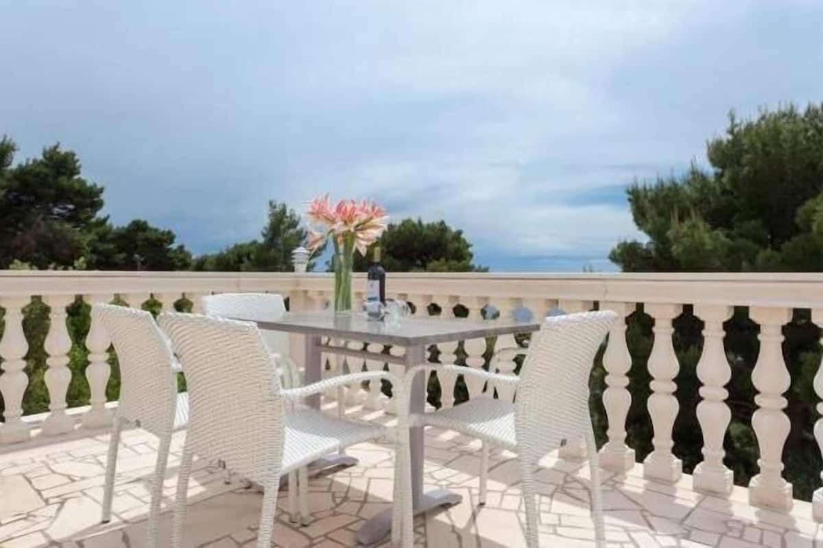 Un balcón con una mesa y sillas, un florero con flores, y vistas a árboles y al cielo.