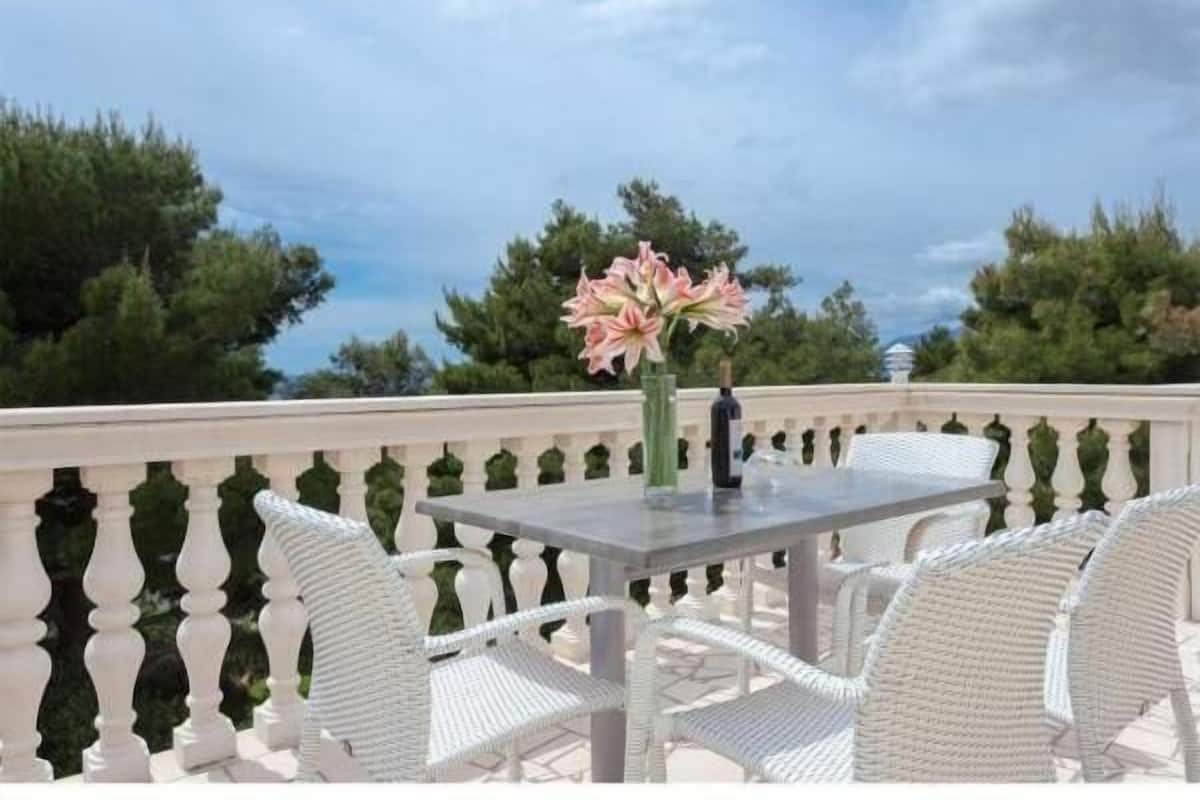 Un balcón con una mesa y sillas, un jarrón con flores, y vistas a árboles y un edificio al fondo.