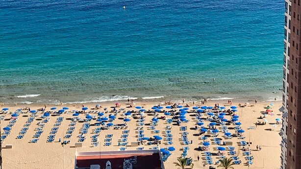 A beach with rows of blue umbrellas and sun loungers, a clear blue sea, and a rocky outcrop in the distance.