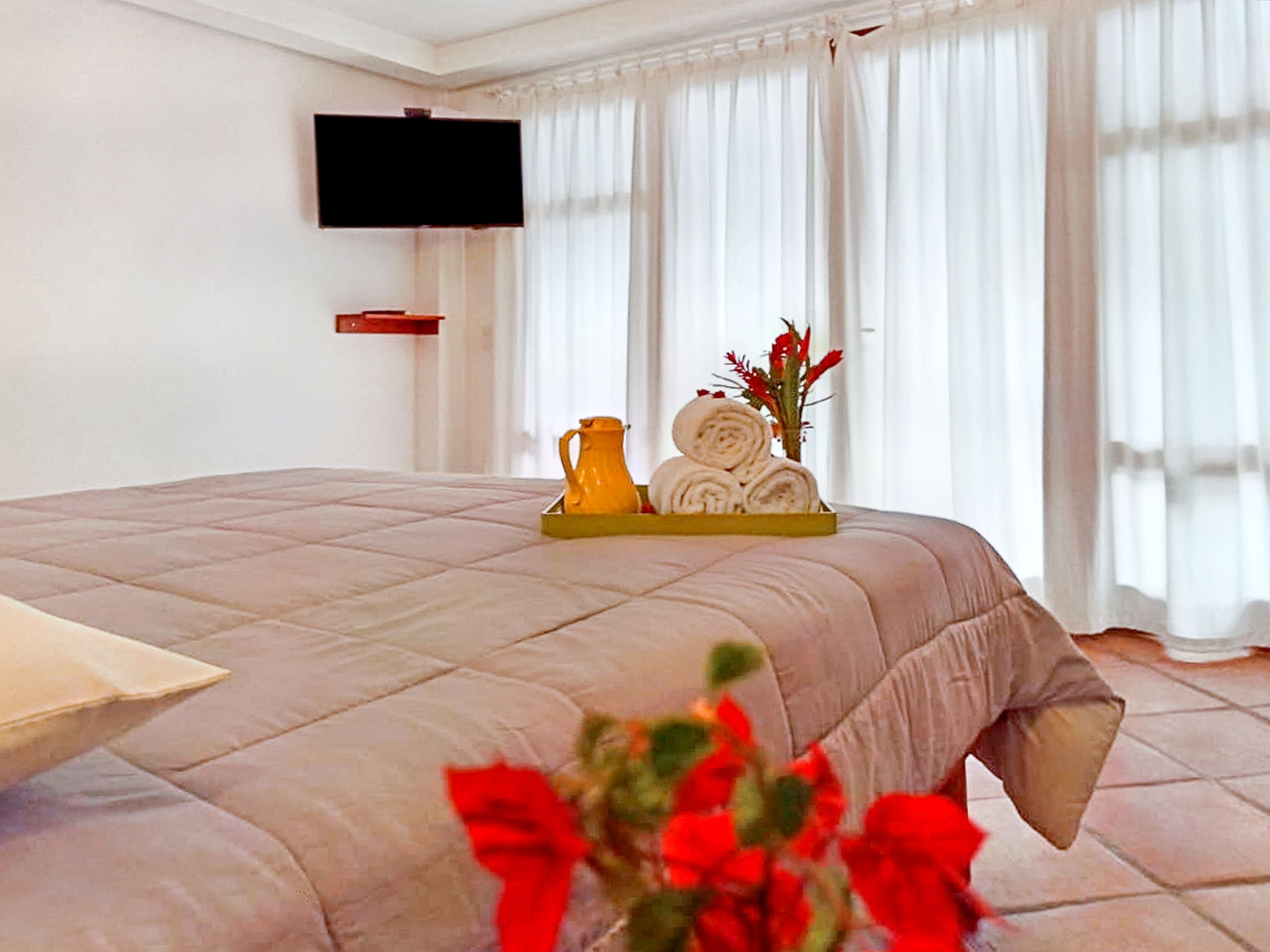 Ein ordentlich bezogenes Bett mit einer beigen Tagesdecke, ein gelber Tablett mit zusammengerollten Handtüchern und eine Vase mit roten Blumen, sowie ein flach an der Wand befestigter Fernseher.