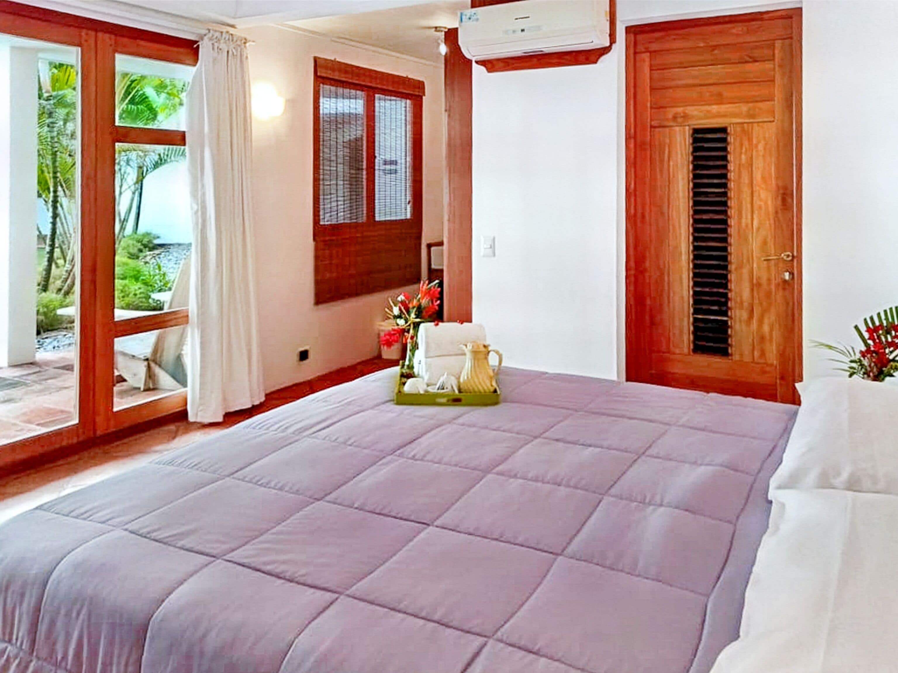 Une chambre avec un grand lit, une fenêtre donnant sur des palmiers, une porte en bois et une petite table sur laquelle est posé un vase de fleurs.