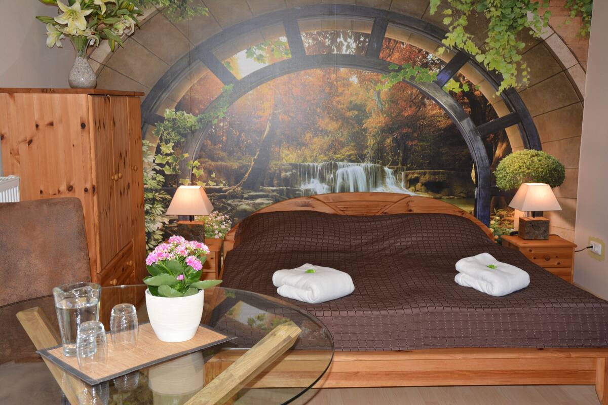 A bedroom with a large bed, two bedside lamps, a glass table with a tray of glasses, a vase with flowers, and a mural of a waterfall.