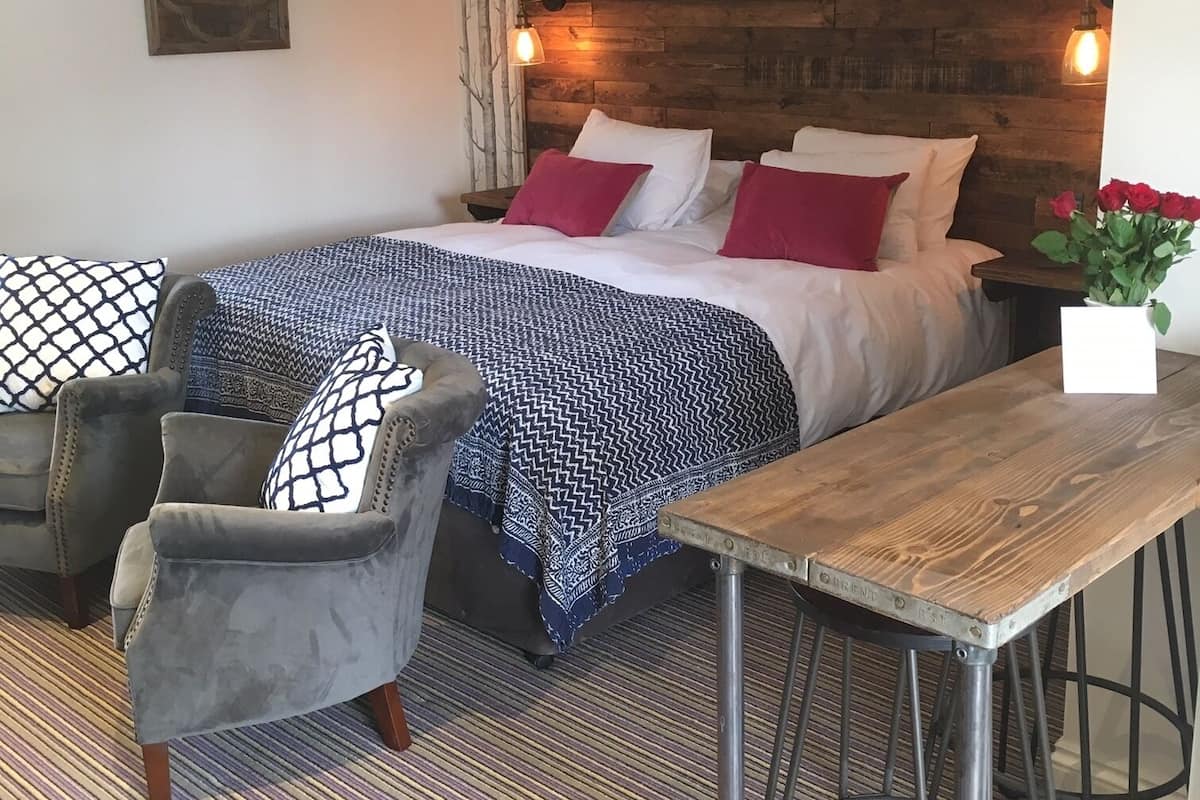 A bedroom with a bed, two armchairs, a wooden table, and a wall-mounted light fixture.