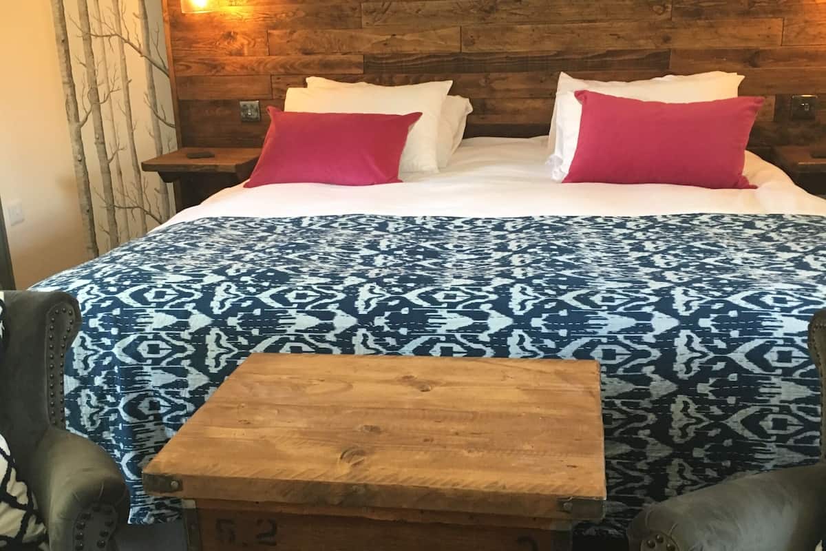 A wooden bed with white and red pillows, a patterned bedspread, and a nightstand with a lamp. There is a small wooden table in front of the bed.