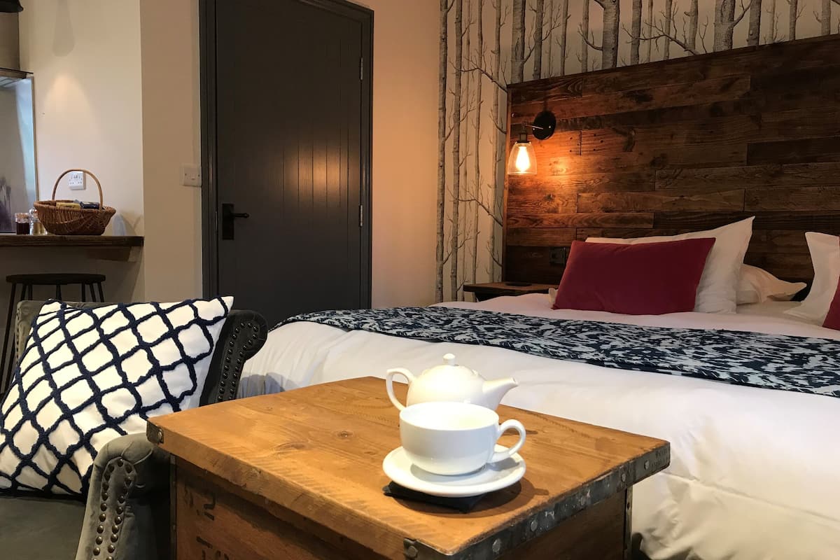 A neatly arranged hotel room with a bed, a wooden nightstand with a teapot, a chair, and a wall with a tree mural.