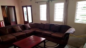 A living room with a sectional sofa, a coffee table, and a patterned armchair. There are two windows with curtains, a door, and a small table in the corner. - Zuus Lodge & Tours (Kalomo)