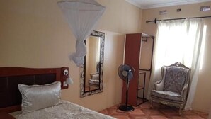 A hotel room with a bed, a chair, a desk, and a fan. - Zuus Lodge & Tours (Kalomo)