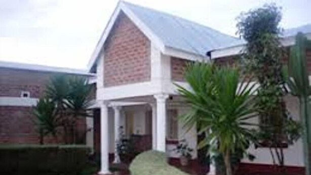 A building with a brick facade and a white porch. There are palm trees and a hedge in front. - Zuus Lodge & Tours (Kalomo)