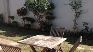 A wooden outdoor table and chairs set on a grassy area with a wall and plants in the background. - Zuus Lodge & Tours (Kalomo)
