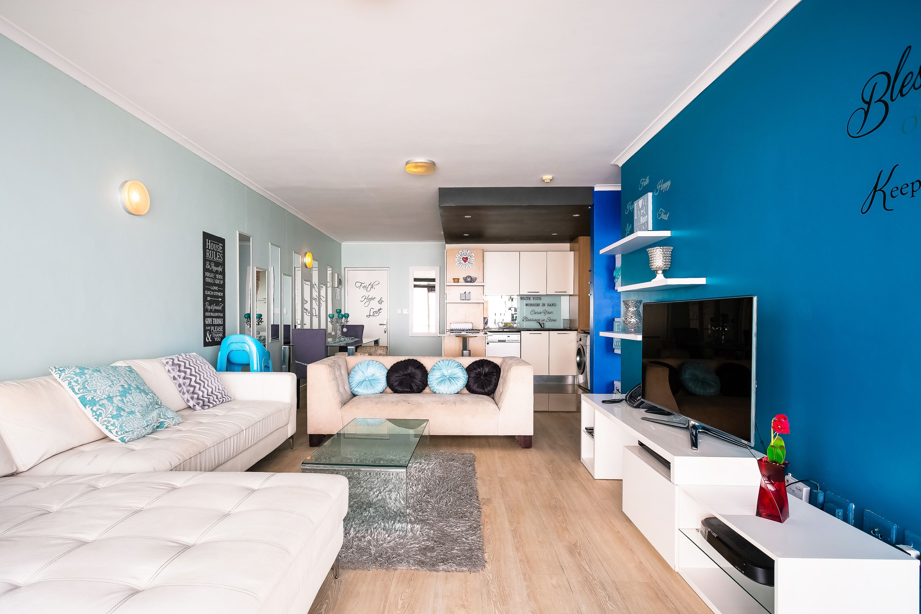 A modern living room with a sofa, coffee table, and a TV on a stand. There is a kitchen in the background with white cabinets and a blue accent wall.