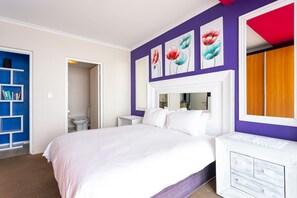 A bedroom with a large bed, a white dresser, and a purple accent wall with floral artwork.