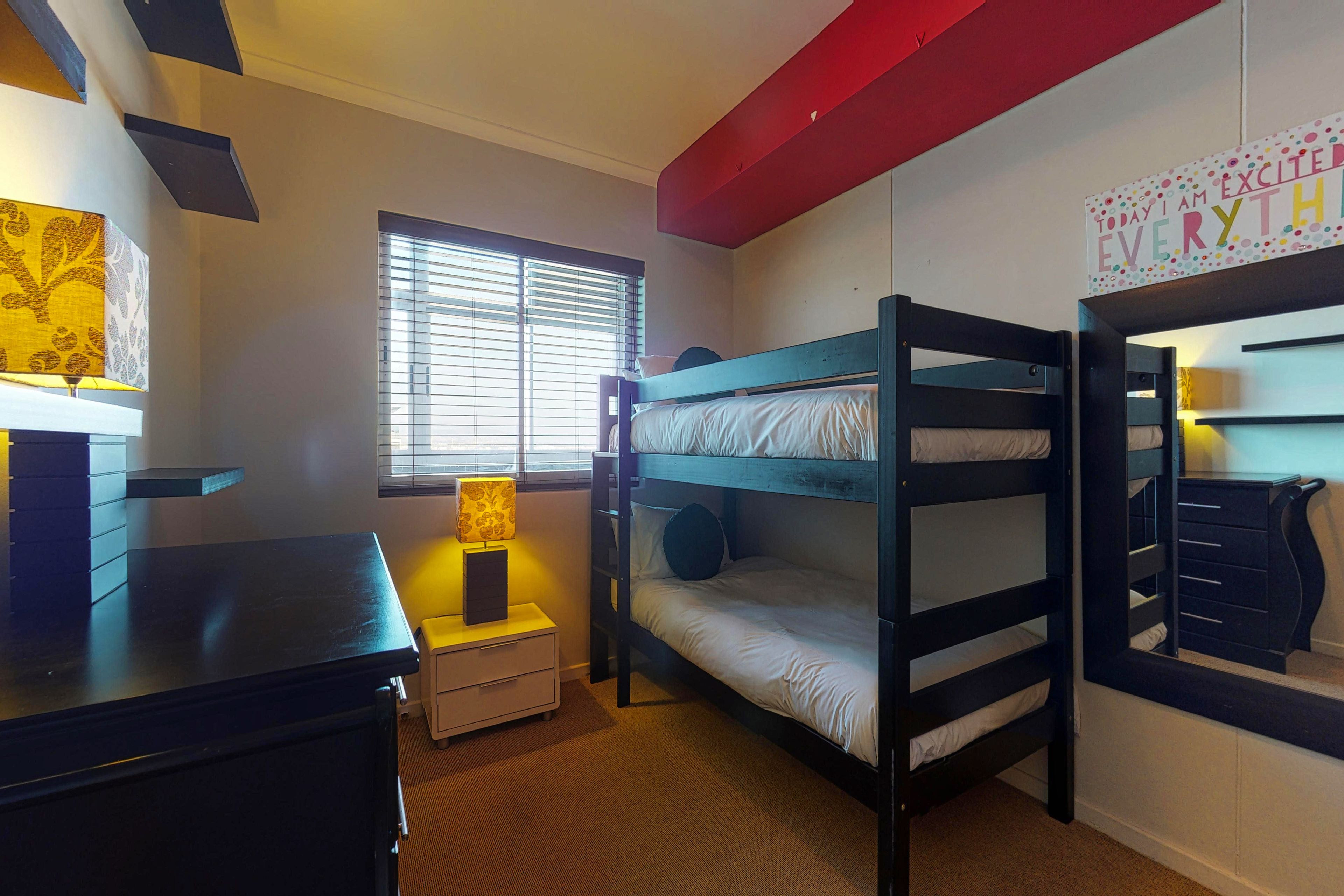 Habitación con literas, un escritorio, una mesita de noche pequeña, una lámpara y una ventana con persianas.