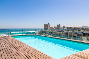 A swimming pool with a wooden deck, overlooking a beach and cityscape.