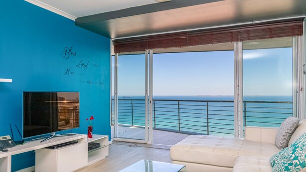 A modern living room with a large TV, a white sofa, and a glass coffee table. There is a balcony with a view of the ocean. - Horizon Breeze 1102 (Cape Town)