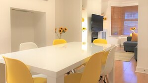 A dining area with a white table and yellow chairs, a painting on the wall, and a TV in the background. - Seaside House With Parking (Worthing)
