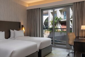 A hotel room with two beds, a desk, and a view of a balcony with plants.