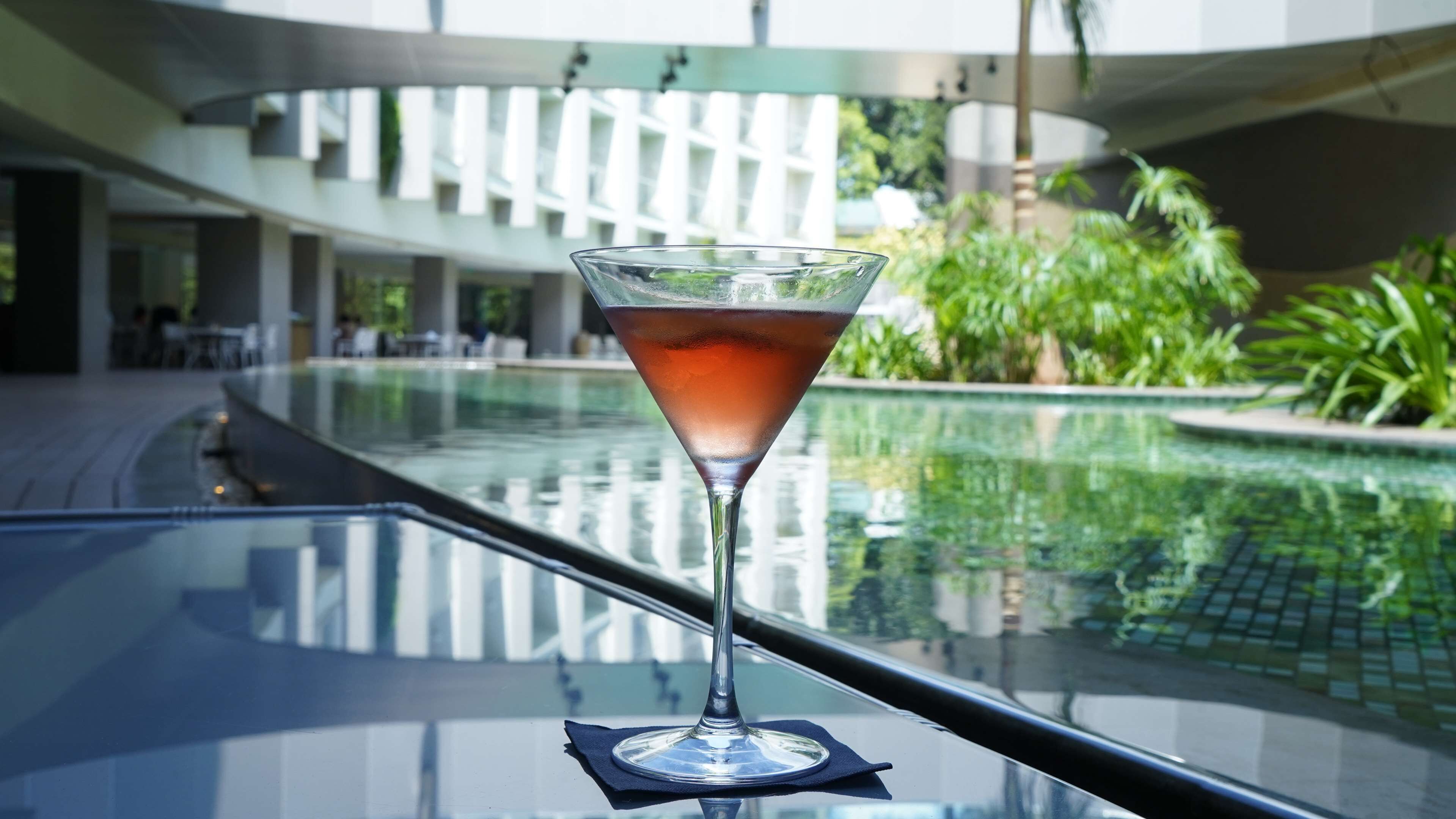 A martini glass on a reflective surface near a pool with a modern building in the background.
