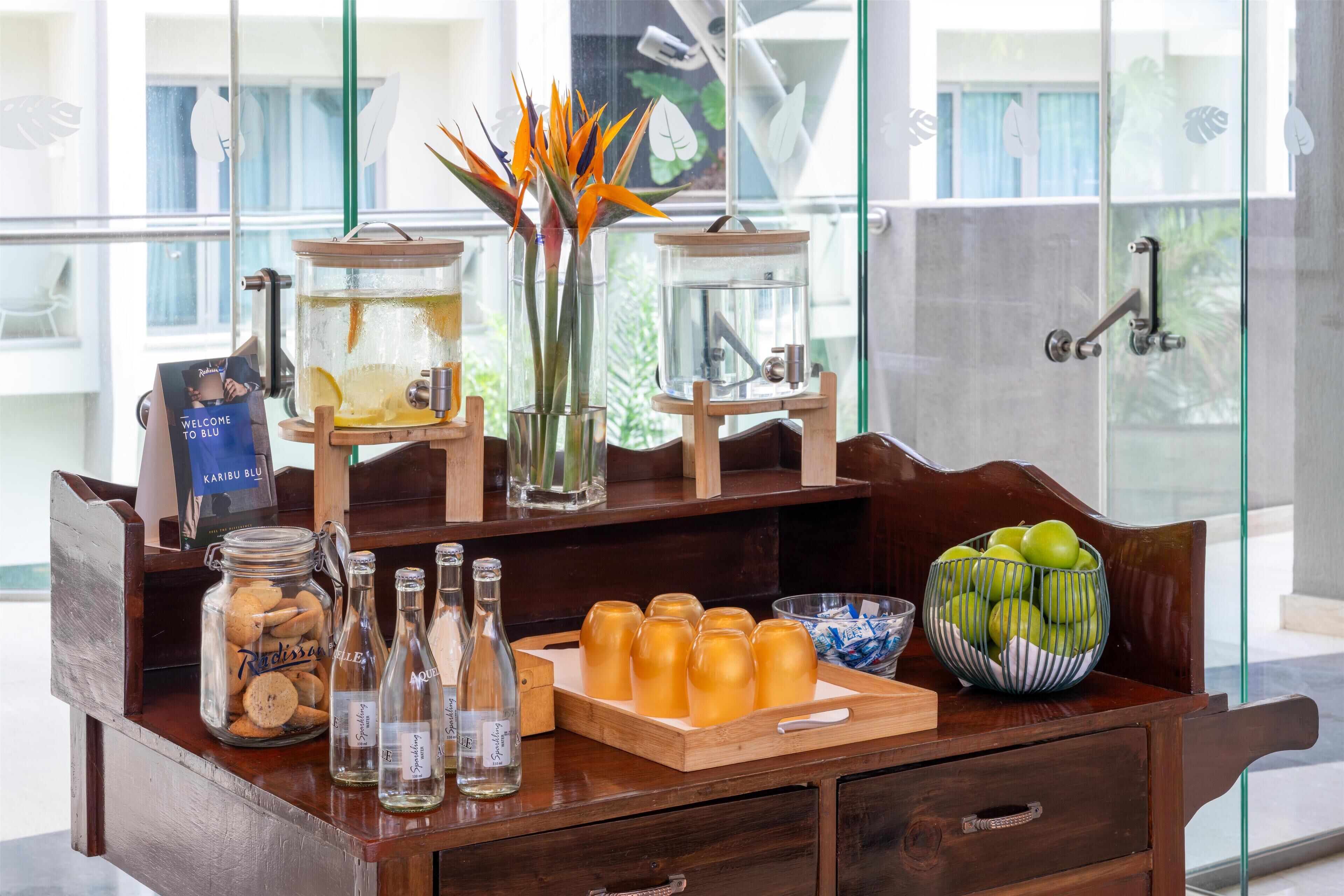 A wooden table with jars of cookies, bottles of water, a tray of glasses, a vase with flowers, and a bowl of apples.