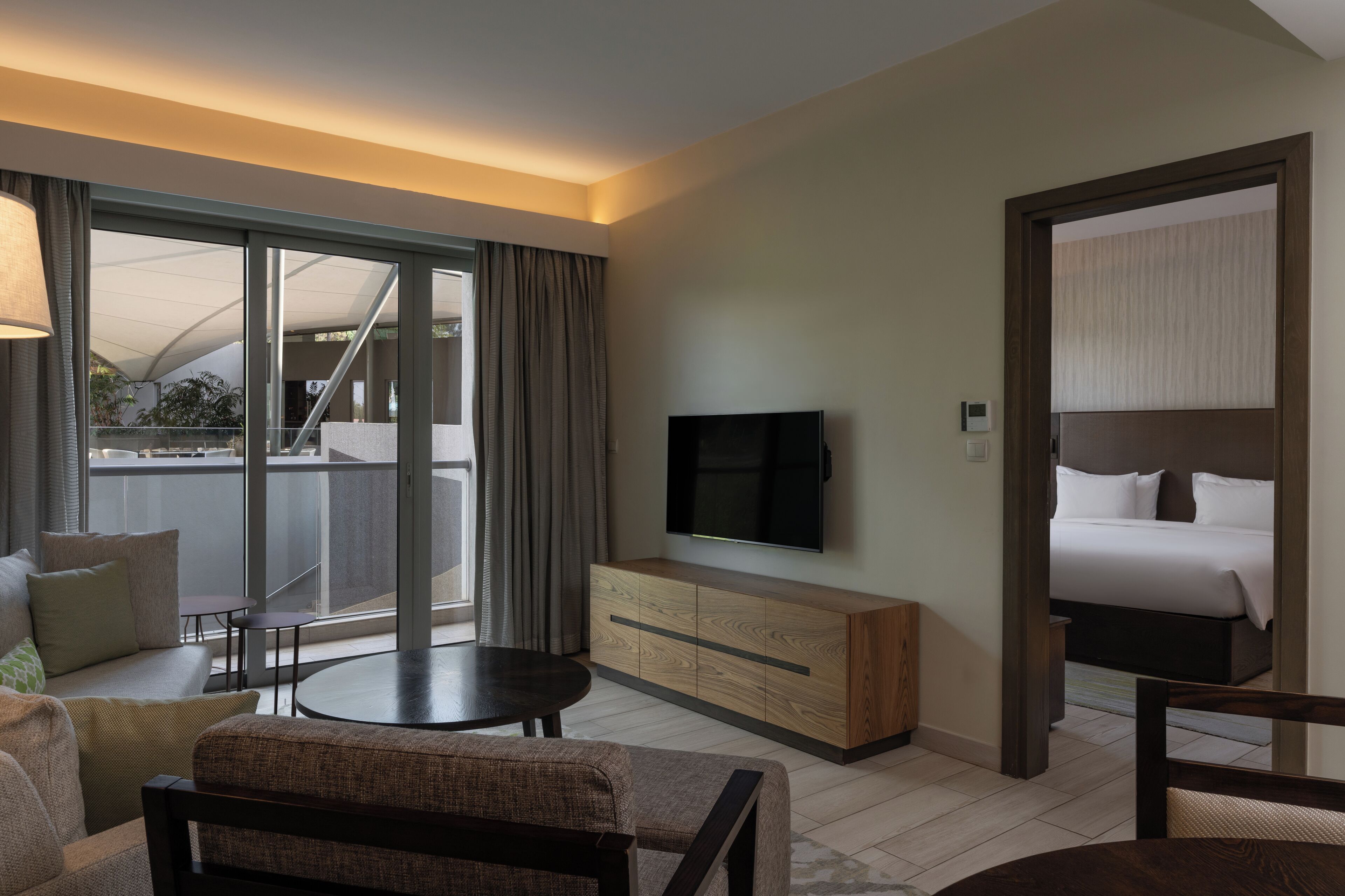 Ein Hotelzimmer mit einer großen Schiebeglasdoor zu einem Balkon, einem wandmontierten Flachbildfernseher, einem hölzernen Konsolentisch, einem runden Couchtisch, einer Couch und einem Bett mit weißen Bettwäsche.